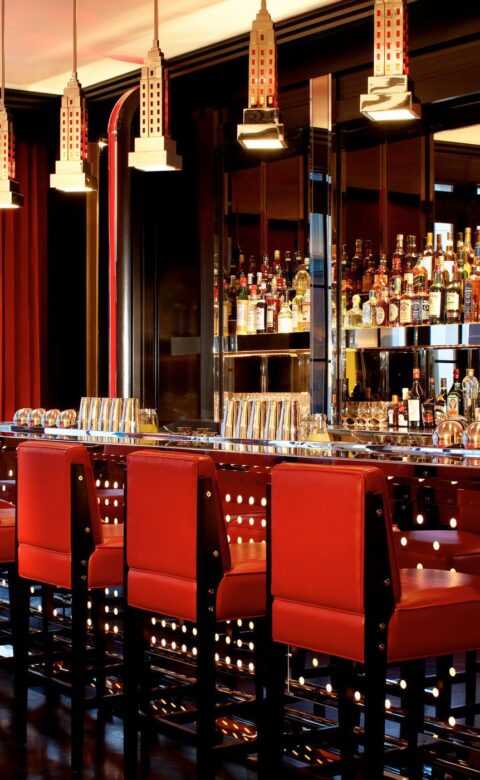 A dark richly colored bar with 6 red chairs and many bottles of liquor