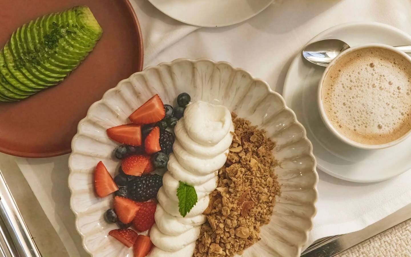 a breakfast of avo slices on one plate, muesli, yoghurt, and berries on another and a cappuccino