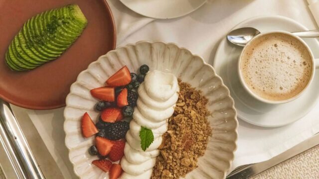 a breakfast of avo slices on one plate, muesli, yoghurt, and berries on another and a cappuccino